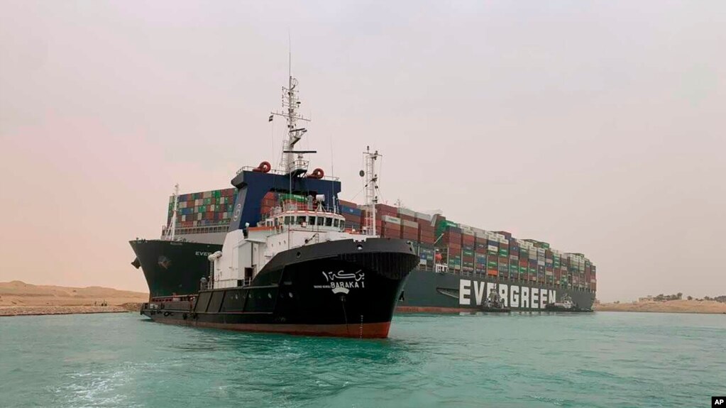 In this photo released by the Suez Canal Authority, a boat navigates in front of a massive cargo ship, named the Ever Green, rear, sits grounded Wednesday, March 24, 2021, after it turned sideways in Egypt’s Suez Canal, blocking traffic in a crucial…