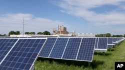 A solar panel array collects sunlight with the Fremont, Neb., power plant seen behind it, May 31, 2018. Solar energy is gaining traction in a small but growing number of Nebraska cities, but the technology still faces a number of obstacles that is keeping