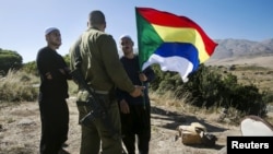FILE - A member of the Druze community holds a Druze flag as he speaks to an Israeli soldier near the border fence between Syria and the Israeli-occupied Golan Heights, near Majdal Shams, June 18, 2015.