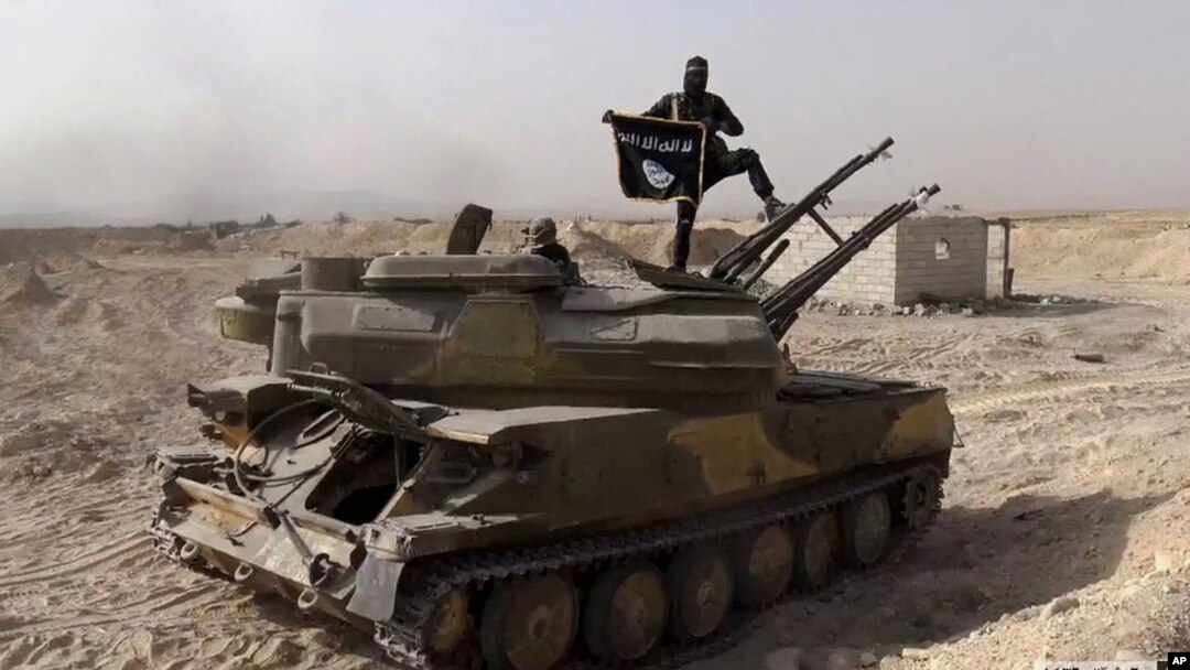 FILE - An Islamic State militant holds the group's flag as he stands on a tank captured from Syrian government forces, in the town of al-Qaryatayn southwest of Palmyra, central Syria. 