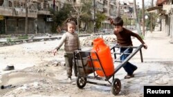 Anak-anak mendorong gerobak berisi wadah air sepanjang jalan yang hancur di kota tua Aleppo, Suriah (11/3). 