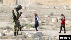 file - A Syrian army soldier looks at his mobile as people visit the ruins of the historic city of Palmyra ahead of a musical event at its amphitheater, Syria, May 6, 2016.