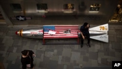 In this Thursday, July 5, 2018, photo, a man sits on a promotional gimmick in the form of a bomb and the U.S. flag outside a U.S. apparel shop at a shopping mall in Beijing, China.