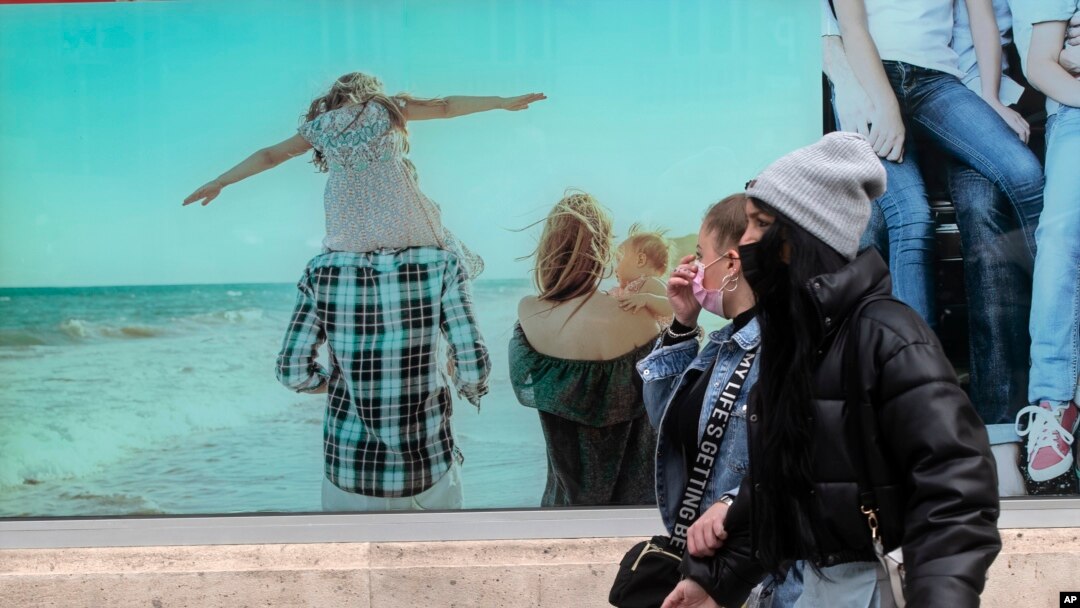 Dua pejalan kaki mengenakan masker untuk melindungi dari penularan virus corona berjalan melintas di depan poster pariwisata di Madrid, Spanyol, 3 Maret 2021. (Foto: AP/Paul White)