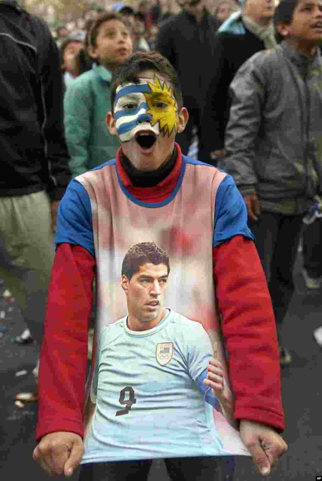 A fan, with his face painted to represent his country's flag and wearing a T-shirt with an image of Uruguayan footballer Luis Suarez, cheers as he watches a live broadcast of the World Cup match between England and Uruguay in downtown Montevideo, Uruguay,
