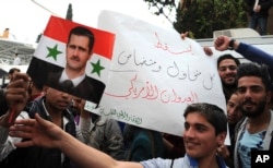 In this photo released by the Syrian official news agency SANA, pro-government protesters hold a portrait of President Bashar Assad and a placard that reads, "Down with everyone who cooperated and supported the American aggression," during a protest against the U.S. attack on a military airbase last week, in front the the U.N. building, in Damascus, April 11, 2017.