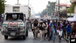 Police officers and protesters take cover behind a police truck as shots ring out during a protest to demand the resignation of Haiti's president Jovenel Moise on the 216th anniversary of the Battle of Vertieres in Port-au-Prince, Haiti, Nov. 18, 2019. 