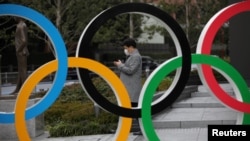 FILE - A man wearing a protective face mask, following the outbreak of the coronavirus, looks at his mobile phone next to the Olympic rings in front of the Japan Olympics Museum in Tokyo, Japan, March 4, 2020. 