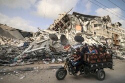 Seorang pria Palestina mengangkut anak-anak dengan sepeda roda tiga melewati gedung Al-Shuruq, yang dihancurkan oleh serangan udara Israel, di Kota Gaza pada 21 Mei 2021, setelah gencatan senjata. (Foto: AFP)