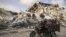 Një burrë palestinez transporton fëmijët ndërsa në sfond duket një godinë e shkatërruar nga sulmet ajrore izraelite (21 maj 2021)