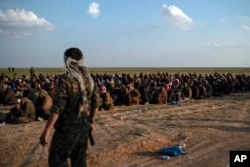 FILE - In this Feb. 22, 2019 file photo, U.S.-backed Syrian Democratic Forces fighters stand guard next to men waiting to be screened after being evacuated out of the last territory held by Islamic State group militants, near Baghuz, eastern Syria.