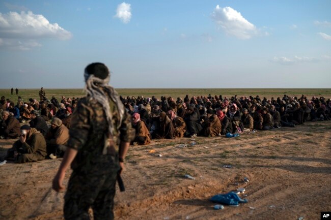 FILE - In this Feb. 22, 2019 file photo, U.S.-backed Syrian Democratic Forces fighters stand guard next to men waiting to be screened after being evacuated out of the last territory held by Islamic State group militants, near Baghuz, eastern Syria.