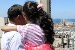 FILE - Makhoul Mohammad al-Hamad stands holding his six-year-old daughter Sama from their apartment as they watch diggers removing earth at the blast site next to the silos at the port of Beirut on Aug. 16, 2020.