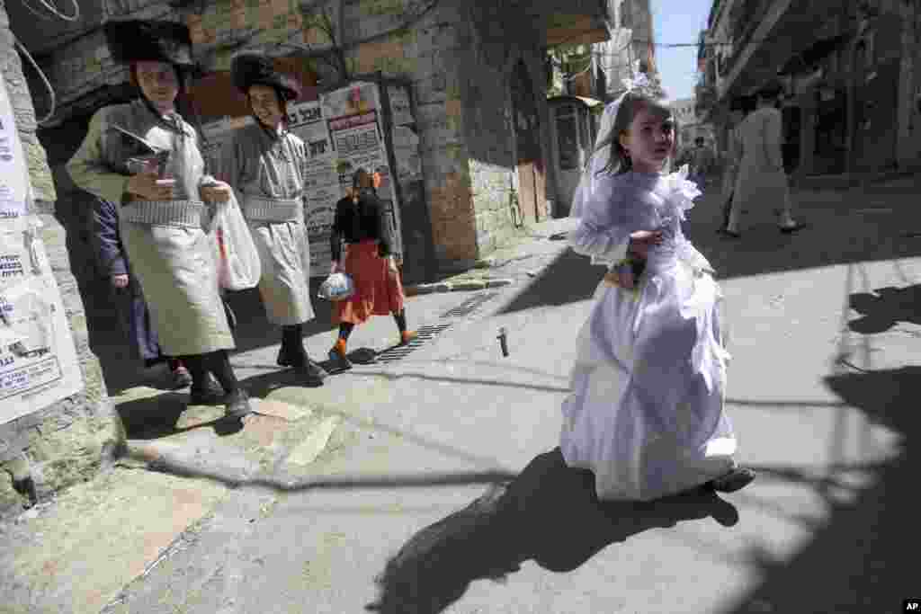 Anak-anak muda pemeluk Yahudi Ortodoks berjalan di samping seorang anak perempuan yang mengenakan kostum pada upacara perayaan Purim di Yerusalem. Festival ini memperingati penyelamatan warga Yahudi dari genosida di Persia seperti ditulis dalam Kitab Ester.