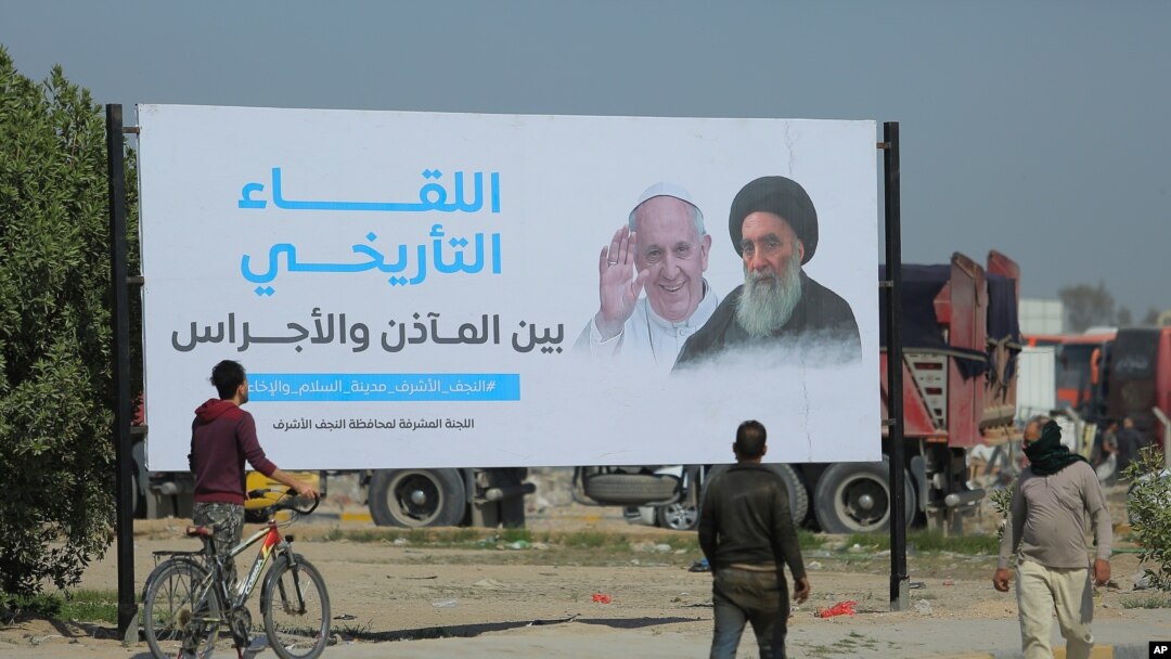 Iraqis walk by a poster announcing the upcoming visit of Pope Francis and his planned meeting with prominent Shiite Muslim leader, Ayatollah Ali al-Sistani, in Najaf, Iraq, March 3, 2021.