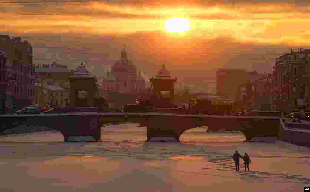A couple walk on the ice on the frozen Fontanka River at sunset in St. Petersburg, Russia, with Troitsky (Trinity) Cathedral in the background.