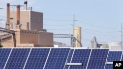 FILE - A solar panel array collects sunlight, in Fremont, Nebraska, May 31, 2018, with a power plant seen behind it.