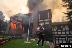 A person carrying a flag walks away from a burning house as powerful winds fueling devastating wildfires in the Los Angeles area force people to evacuate in Altadena, California, Jan. 8, 2025.