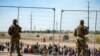 ARCHIVO - Varios migrantes esperan junto a la cerca fronteriza ante la mirada de guardias nacionales de Texas para entrar en El Paso, Texas, el miércoles 10 de mayo de 2023.