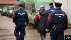 FILE - A migrant is seen escorted past police vehicles at a detention center for illegal migrants, in Szeged, southern Hungary, Dec. 17, 2014.