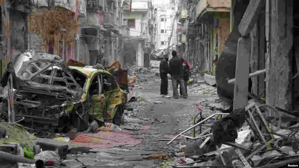 Civilians stand between damaged buildings and vehicles in the besieged area of Homs, Jan. 27, 2014.