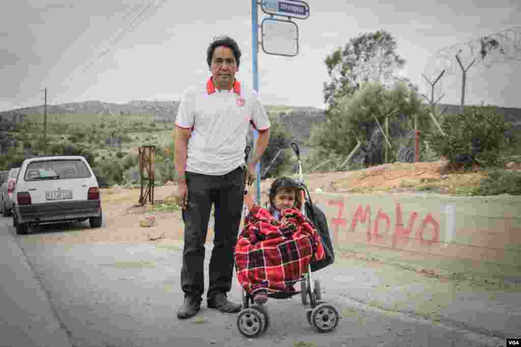 Seorang ayah dan anak perempuannya menunggu bus di luar kamp Schisto, yang terletak beberapa kilometer dari pusat kota Athena. Beberapa kamp lain jauh lebih terisolasi. (VOA/J. Owens)