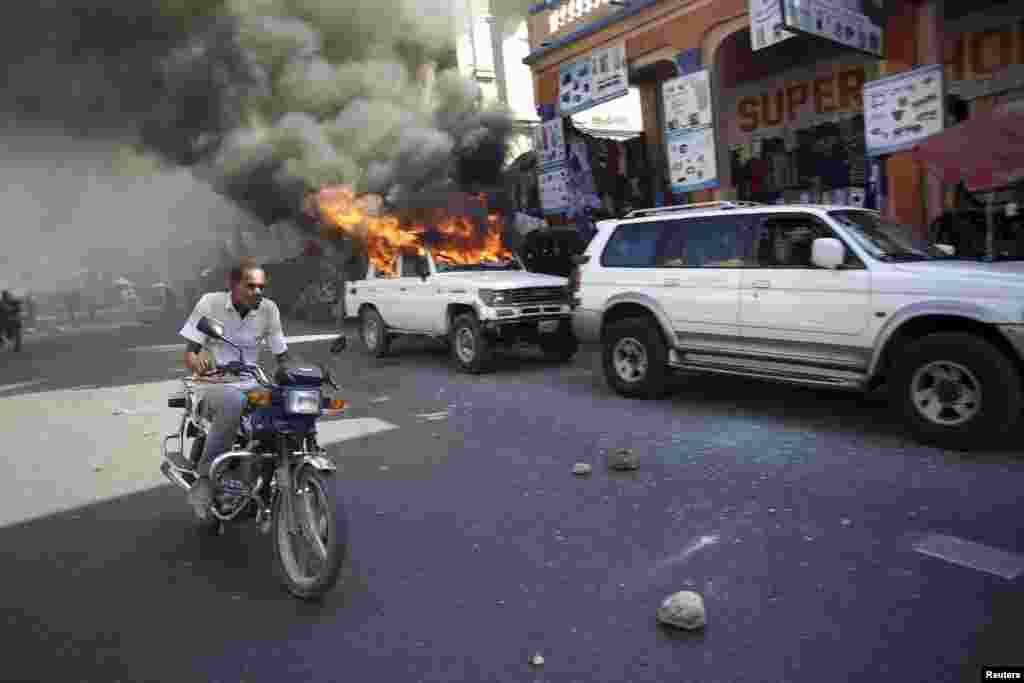 A man on a motorcycle passes next to a car that was set afire by protesters during a demonstration against the electoral process in Port-au-Prince, Haiti, Jan. 18, 2016.