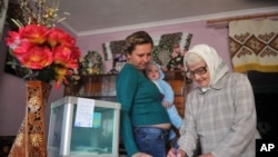 An elderly woman fills in the ballot while a woman with a child watches, as they vote at home in the village of Stilske, west Ukraine on Sunday, Oct. 25, 2015. 