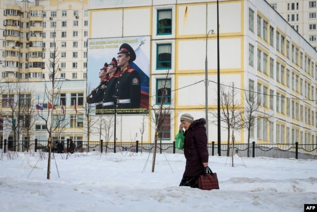FILE - A woman carrying bags of groceries walks past a school building with a military-themed poster in front of it, on the outskirts of Moscow, Russia, Feb. 27, 2019.