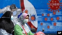 Dua orang perempuan sedang mengendarai sepeda motor melewati mural bertema virus corona di Jakarta, 23 Desember 2020. (AP Photo)