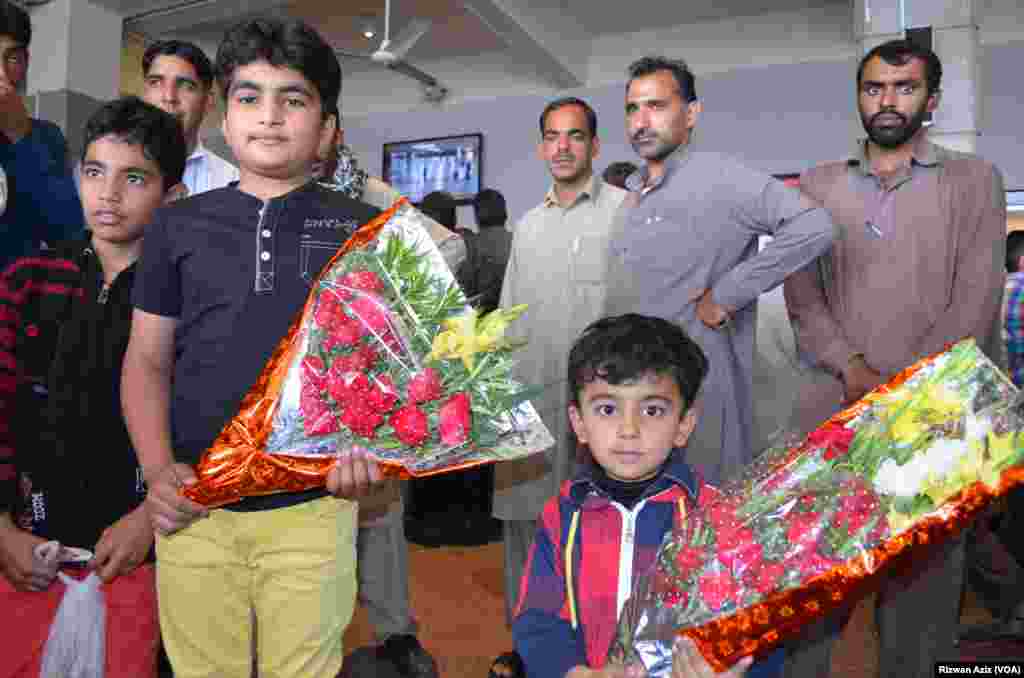 دو بچے اپنے عزیز کے استقبال کے لیے پھول لے کر کھڑے ہیں۔