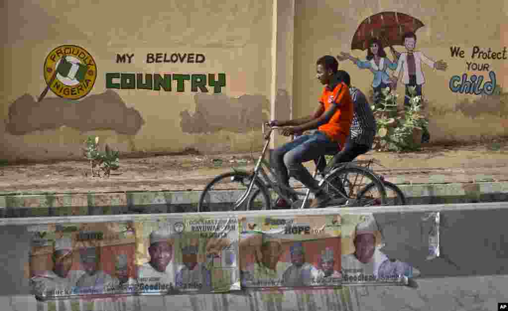 Dois jovens andando de bicicleta na frente de um mural na parede de uma escola e pôsteres eleitorais a favor do presidente Goodluck Johnathan em Kano.