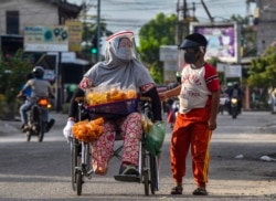 Seorang perempuan berkursi roda dan putranya, mengenakan masker dan pelindung plastik di tengah pandemi COVID-19, berjualan jajanan di sepanjang jalan di Pekanbaru, Riau, 20 Juni 2020. (Foto: WAHYUDI/AFP)