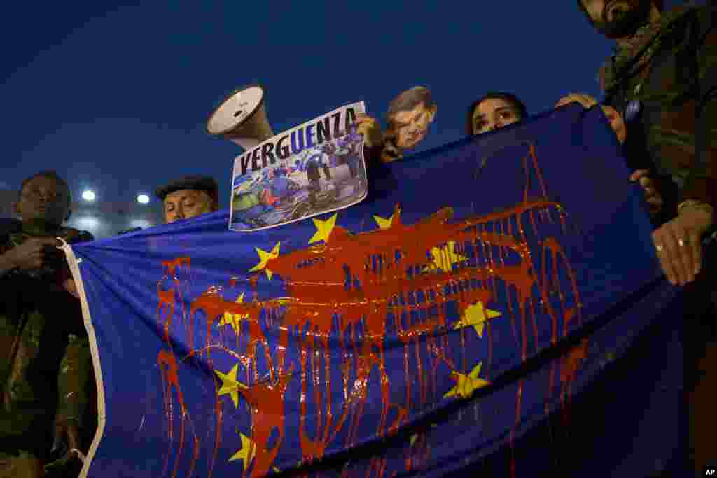People hold a European Union flag stained with red paint as they protest in Madrid, Spain, against a possible deal between the EU and Turkey to return thousands of migrants. The poster shows a photograph of a refugee camp and reads in Spanish: &quot;Shame.&quot;