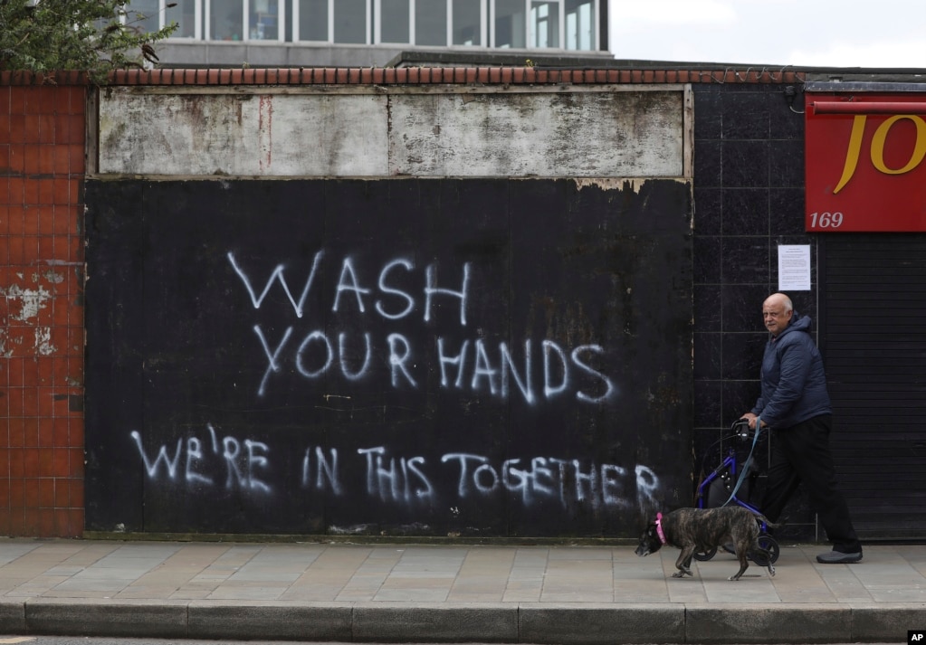 Un hombre pasa a su perro cerca de una pared con un graffiti que pide a las personas que se laven las manos, en Belfast, Irlanda del Norte.