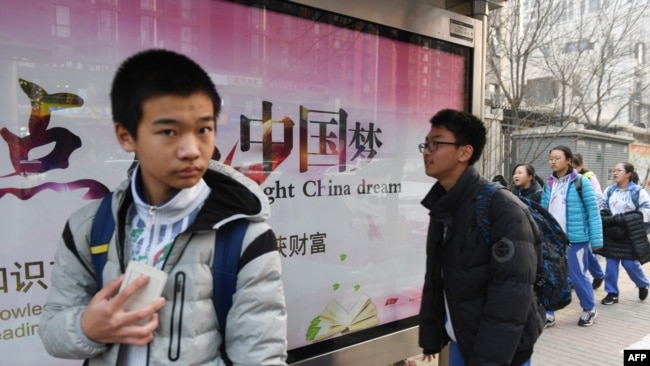 2018年3月12日，北京一所学校外面，青少年走过宣传牌，牌上写着中英文口号“点亮中国梦 知识改变命运 付出收获财富”。