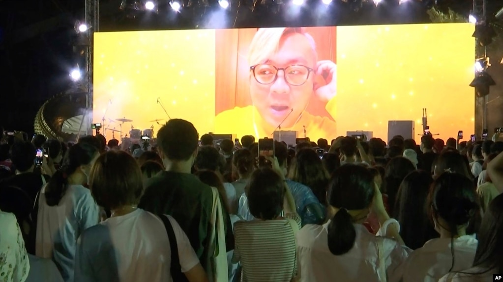 In this image taken from video, Pavin Chachavalpongpun, administrator of the Facebook group “Royalist Marketplace,” is shown delivering a pre-recorded message onscreen before a crowd attending an anti-government rally on Aug. 10, 2020, at Thammasat University in Thailand.