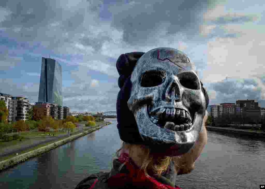 A young man wears a mask on the right side of his head as he stands on a bridge with the European Central Bank at left in Frankfurt, Germany.