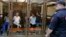 Defendants in the murder trial of Russian investigative journalist Anna Politkovskaya are seen inside a glass-walled cage, with a policeman standing guard in the foreground, during a court hearing in Moscow, June 9, 2014.