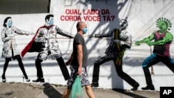 រូបឯកសារ៖ បុរសម្នាក់ពាក់ម៉ាស់ដើរកាត់រូបលើជញ្ជាំងមួយបង្ហាញពីការប្រទាញប្រទង់គ្នារវាងបុគ្គលិកសុខាភិបាល និងប្រធានាធិបតីលោក Jair Bolsonaro ដែលត្រូវបានជួយដោយរូបតុក្កតាវីរុសកូរ់ូណា នៅក្រុង Sao Paulo ប្រទេសប្រេស៊ីល កាលពីថ្ងៃទី១៩ ខែមិថុនា ឆ្នាំ២០២០។