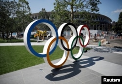 FILE - Construction workers and passersby are seen through Olympic rings in front of the construction site of the New National Stadium, the main stadium of Tokyo 2020 Olympics and Paralympics, in Tokyo, Japan, June 13, 2019.