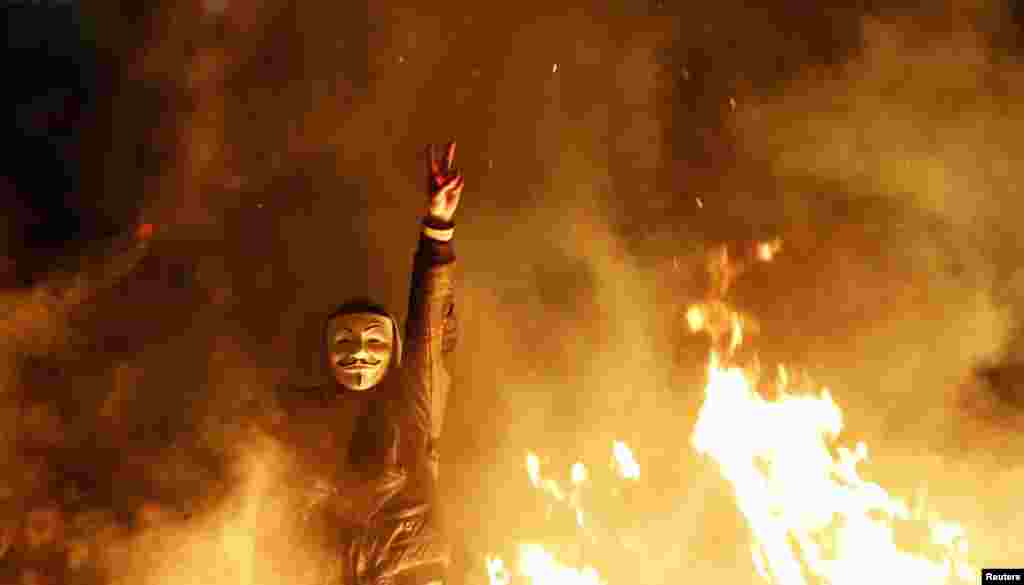 An anti-government protester wearing a Guy Fawkes mask gestures behind a barricade that they set on fire during a demonstration in Ankara, Turkey, Mar. 12, 2014. Riot police clashed with demonstrators in several Turkish cities for a second day as mourners buried a teenager wounded in protests last summer.