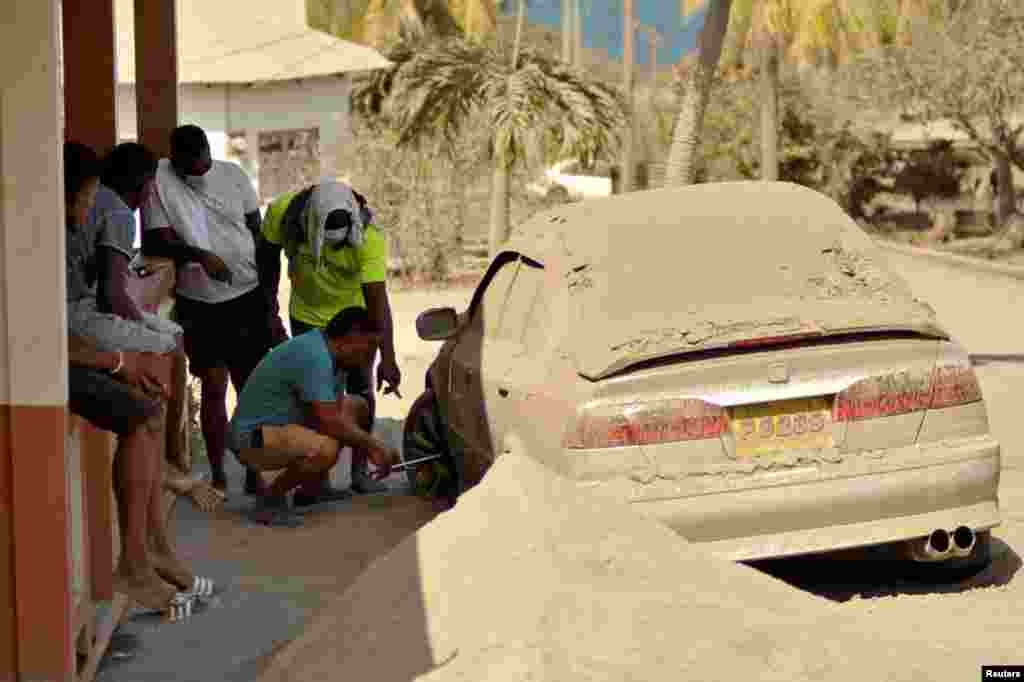 A man fixes a flat tire of a car covered in ash after a series of eruptions from La Soufriere volcano in Orange Hill, Saint Vincent and the Grenadines, April 18, 2021.