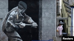 FILE - A woman takes out her rubbish beside a mural depicting a British soldier knocking down the door of a house with a sledge hammer in Derry, Northern Ireland, March 23, 2010. The Bloody Sunday massacre took place in Derry in 1972.