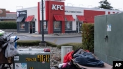 Una persona sale de un restaurante de la cadena de comida rápida KFC mientras otra persona sin hogar duerme en la concurrida intersección de Oregon Coast Highway y la 5th Street el 19 de junio de 2024, en Brookings, Oregon, EEUU.