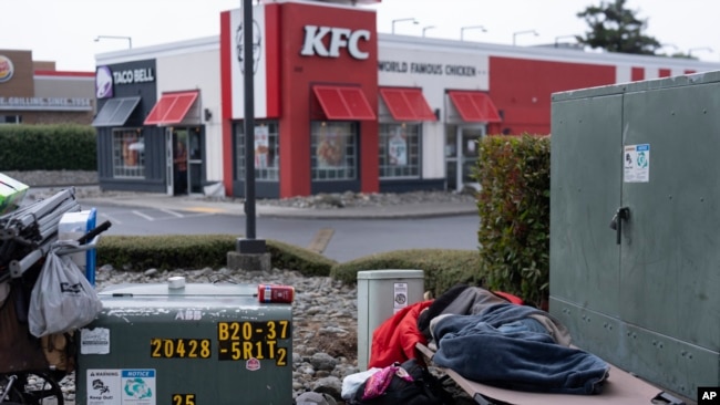 Una persona sale de un restaurante de la cadena de comida rápida KFC mientras otra persona sin hogar duerme en la concurrida intersección de Oregon Coast Highway y la 5th Street el 19 de junio de 2024, en Brookings, Oregon, EEUU.
