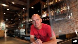 Jeff Brightwell, owner of Dot's Hop House, is framed between protective shielding placed over the bar as he poses for a photo at his establishment in the Deep Ellum entertainment district in Dallas, Aug. 20, 2020.