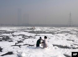 A woman bathes her daughter in the Yamuna River, covered by a chemical foam caused by industrial and domestic pollution as the skyline is enveloped in toxic smog, in New Delhi, India, Nov. 17, 2021.