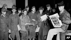 Pfc. Clarence K. Ayers of Evansville, Ind., reads the news of VE Day as newly arrived German prisoners stand of a New York City pier on May 8, 1945.