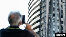 A man takes a picture as firefighters work on the blackened ruins of a block of flats after a fire ripped through the building, in Milan, Italy, Aug. 30, 2021. 
